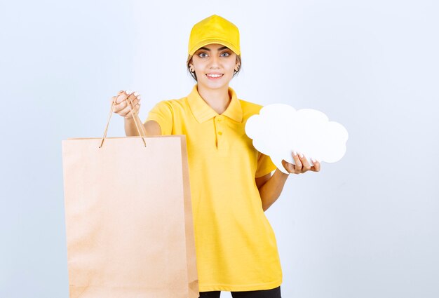 Une jolie femme en uniforme jaune tenant un sac en papier kraft vierge marron et une bulle de dialogue vierge.