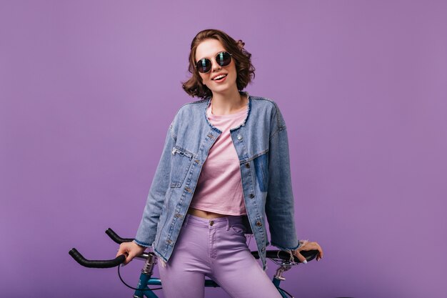 Jolie femme en veste en jean posant avec vélo. Tir à l'intérieur d'une femme bouclée confiante isolée.