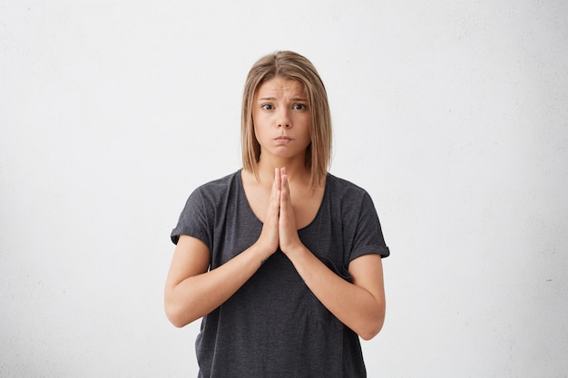 Jolie femme en vêtements décontractés aux yeux noirs et cheveux blonds ayant un regard innocent en appuyant sur ses mains posant ensemble. Regretful belle jeune femme demandant des excuses