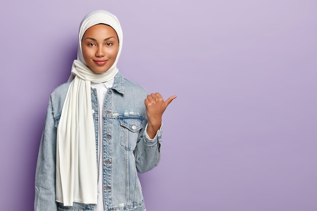 jolie femme en vêtements traditionnels arabes, pointe le pouce vers le côté droit, présente un objet sur un espace vide, a des opinions religieuses, isolé sur un mur violet. Concept de religion