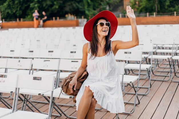 Jolie femme vêtue d'une robe blanche, chapeau rouge, lunettes de soleil assis dans le théâtre en plein air d'été sur une chaise seule, tendance de la mode printemps street style, agitant la main