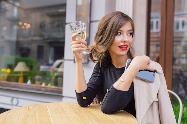 Jolie femme vêtue d'une robe noire et trench beige avec une coiffure élégante et des lèvres rouges sur une terrasse