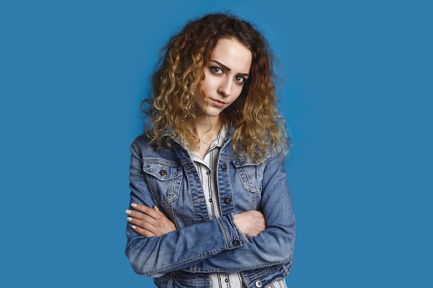 Jolie fille de 20 ans avec des cheveux bouclés lâches posant ayant un regard méfiant suspect, gardant les bras croisés, portant une veste en jean élégante. Expressions faciales humaines, émotions et langage corporel