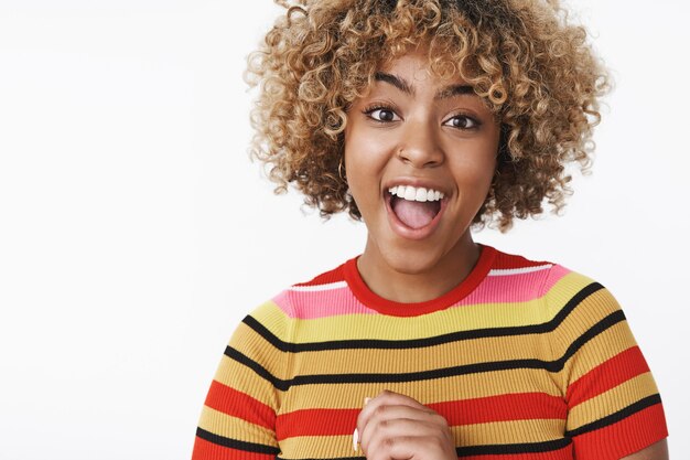 Jolie fille afro-américaine enthousiaste et excitée dans un pull rayé élégant souriant la bouche largement ouverte de frisson et de joie, réagissant à des nouvelles étonnantes, heureuse pour un ami sur un mur blanc