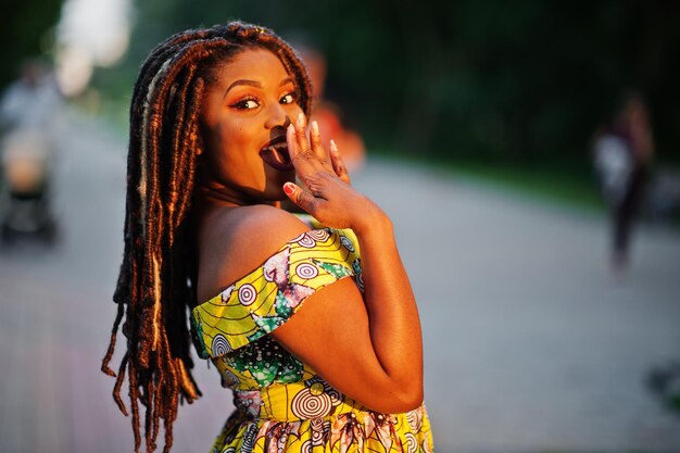 Jolie fille afro-américaine de petite taille avec des dreadlocks porter à la robe jaune colorée posée au coucher du soleil émotion surprise