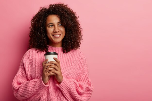 Jolie fille afro tient une tasse de café en papier, aime le temps libre, regarde avec le sourire de côté, porte un pull surdimensionné