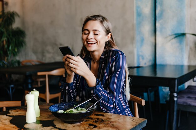 Jolie fille à l'aide de téléphone pendant le dîner au café. Portrait de jeune femme heureuse, manger des légumes.