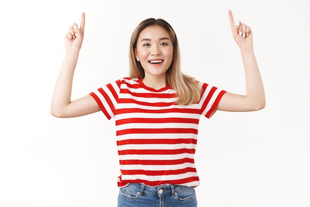 Jolie fille asiatique blonde suggère un voyage de vacances parfait pointant vers le haut l'index agence de voyage promo souriant largement regarder excitation joie caméra debout tshirt rayé rouge fond blanc