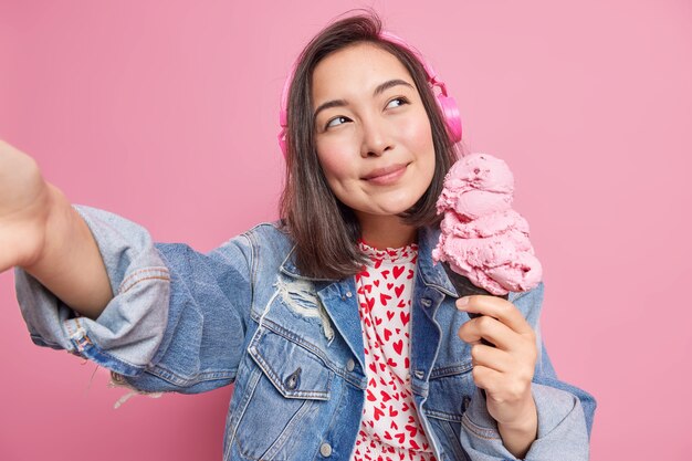 Une jolie fille asiatique brune réfléchie et rêveuse pose avec une grosse glace délicieuse dans une gaufre imagine quelque chose d'agréable fait une photo d'elle-même écoute de la musique via un casque stéréo a des vacances d'été