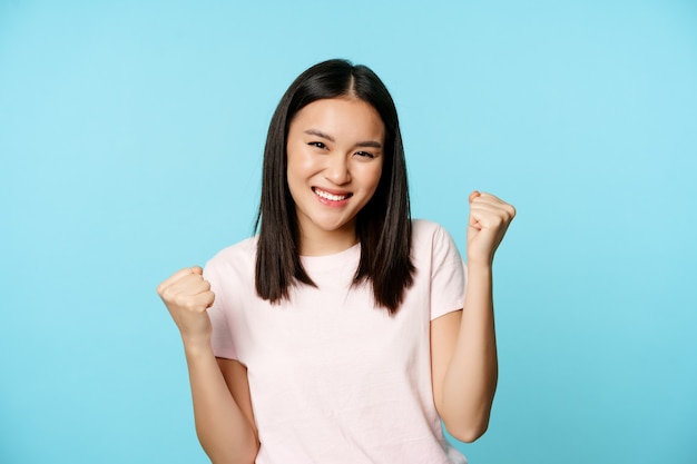 Jolie fille asiatique enthousiaste gagnante, se réjouissant d'une bonne nouvelle, danse de la victoire, souriante heureuse, debout sur fond bleu