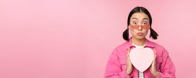 Photo gratuite jolie fille asiatique montrant une boîte en forme de coeur avec un cadeau à la recherche d'un concept de présent romantique surpris et excité portant des lunettes de soleil debout sur fond rose