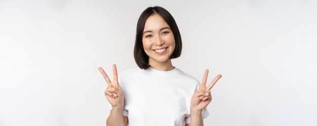 Jolie fille asiatique montrant la paix vsign souriant et regardant heureux à la caméra portant un fond de studio de t-shirt blanc