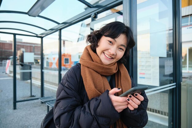 Photo gratuite jolie fille asiatique souriante debout sur l'arrêt de bus tenant un smartphone portant une veste d'hiver et une écharpe woma