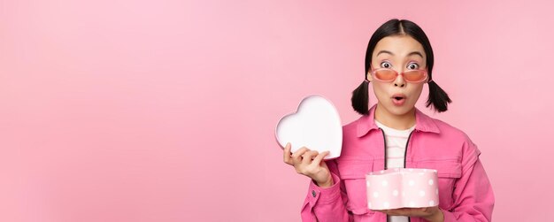 Jolie fille asiatique vous donnant un cadeau dans une boîte en forme de coeur embrassant et souriant concept de vacances et de célébration debout sur fond rose