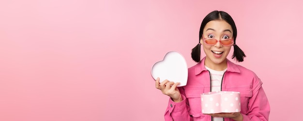 Jolie fille asiatique vous donnant un cadeau dans une boîte en forme de coeur embrassant et souriant concept de vacances et de célébration debout sur fond rose