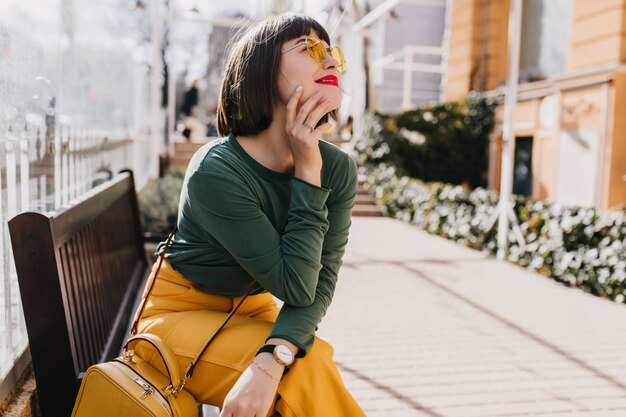 Jolie fille aux cheveux bruns en pull vert se détendre sur un banc. Portrait en plein air d'une femme magnifique à lunettes de soleil rêveuse posant dans la rue.