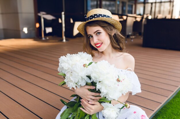 Jolie fille aux cheveux longs au chapeau est assise sur le sol sur la terrasse. Elle porte une robe blanche aux épaules nues, rouge à lèvres rouge. Elle a des fleurs blanches dans les mains et sourit.