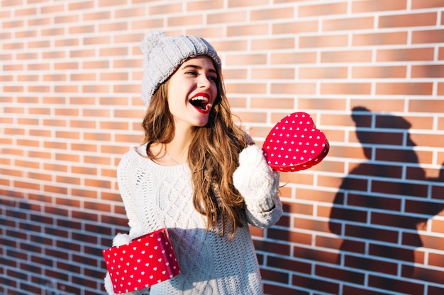 Jolie fille aux cheveux longs en bonnet tricoté et gants sur le mur extérieur. Elle tient le cœur ouvert dans les mains, exprimant sur le côté.