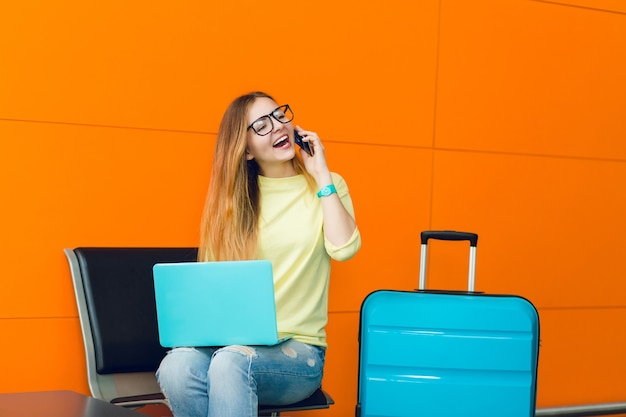 Jolie fille aux cheveux longs est assise sur une chaise sur fond orange. Il y a un ordinateur portable bleu sur ses genoux et une valise bleue à proximité. Elle parle au téléphone.