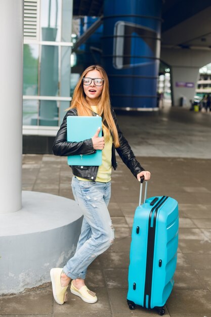 Jolie fille aux cheveux longs est debout avec une valise à l'extérieur à l'aéroport. Elle porte une veste noire avec un jean et tient un ordinateur portable. Elle a l'air surprise.