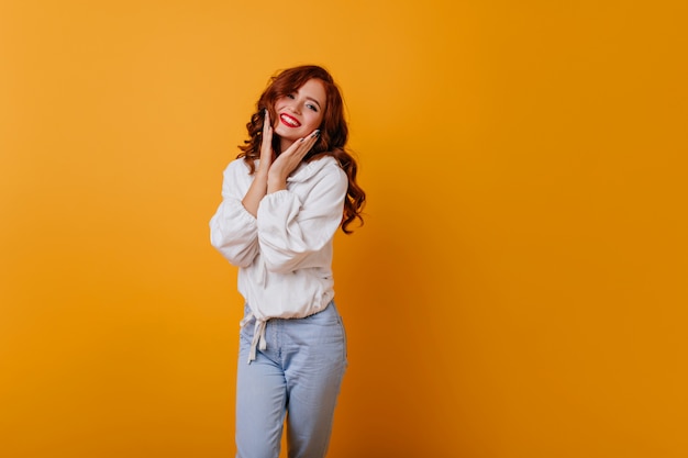 Jolie fille aux cheveux longs posant avec un joli sourire. Gracieuse jeune femme blanche debout sur un mur jaune.