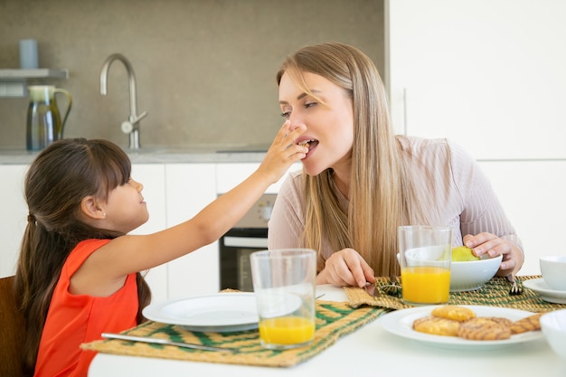 Jolie fille aux cheveux noirs donnant un cookie à sa mère pour la dégustation et la morsure, prenant le petit déjeuner avec sa famille