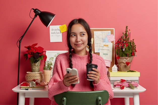 Jolie fille aux cheveux noirs utilise un smartphone pour surfer sur les réseaux sociaux, boit du café à emporter, détourne les yeux, vêtue d'un pull en tricot, pose contre le bureau avec un arbre de Noël décoré