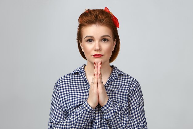 Jolie fille aux cheveux roux et rouge à lèvres en appuyant sur les paumes ensemble dans la prière. Belle jeune femme élégante priant en studio, regardant la caméra avec de grands yeux pleins d'espoir et de forte conviction