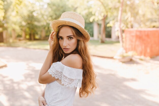 Jolie fille aux longs cheveux blonds posant de manière ludique après une promenade dans le parc. Charmante dame en robe de dentelle et chapeau avec ruban blanc se tenant dehors avec une expression de visage sérieuse.