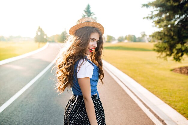 Jolie fille aux longs cheveux bouclés au chapeau marche sur la route au coucher du soleil. Elle porte un T-shirt blanc, un justaucorps, une jupe, des lèvres rouges. Elle a l'air heureuse au soleil.