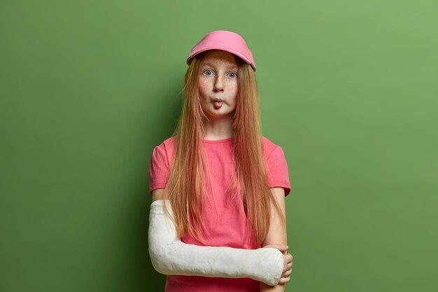 Une jolie fille aux taches de rousseur fait une grimace, des lèvres de poisson, a de longs cheveux roux droits, porte une casquette et un t-shirt roses, un bras cassé en plâtre, se tient contre le mur vert. Enfants, expressions du visage, accident