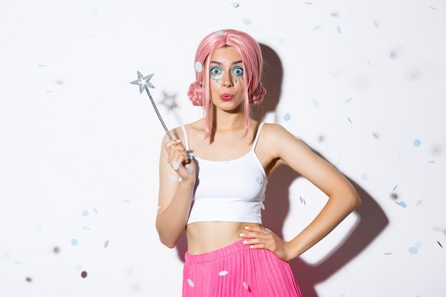 Jolie fille avec baguette magique célébrant l'halloween en costume de fée et perruque rose, regardant coquette à la caméra, debout pendant que les confettis tombent.