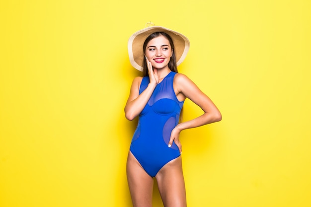 Jolie fille en bikini blanc, chapeau, lunettes de soleil, bouche ouverte émotionnellement sur un mur jaune avec un corps parfait. Isolé.