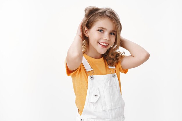 Jolie fille blonde assez heureuse se sentant optimiste, profitez des vacances d'été, sans devoirs, portez un t-shirt jaune et une salopette, touchez les cheveux, souriant heureux
