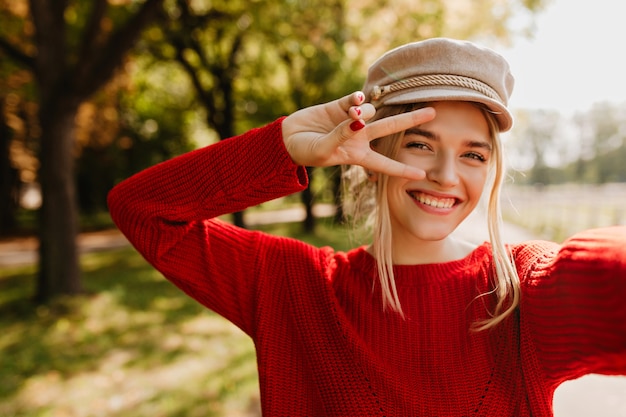 Jolie fille blonde au chapeau élégant et pull rouge posant avec le sourire pour faire un selfie dans le parc.
