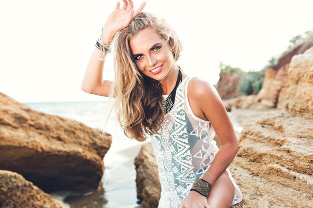 Jolie fille blonde aux cheveux longs est assise sur la pierre sur la plage rocheuse sur fond de coucher de soleil. Elle sourit à la caméra.