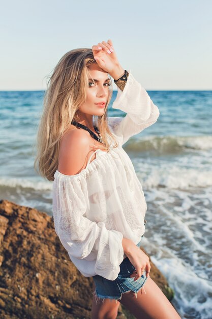 Jolie fille blonde aux cheveux longs pose à la caméra sur fond de mer. Elle porte une chemise blanche avec des épaules nues et un short.