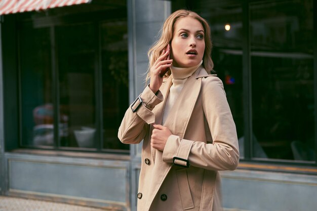 Jolie fille blonde décontractée en trench-coat parlant au téléphone portable regardant rêveusement à l'extérieur