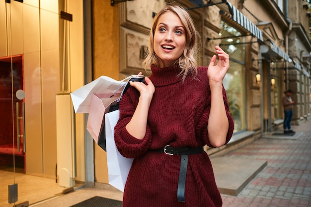 Jolie fille blonde élégante en pull tricoté avec des sacs à provisions regardant joyeusement dans la rue de la ville