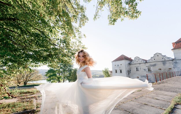 Jolie fille blonde heureuse vêtue d'une robe blanche se retourne et sourit aux beaux jours près du vieux château de pierre