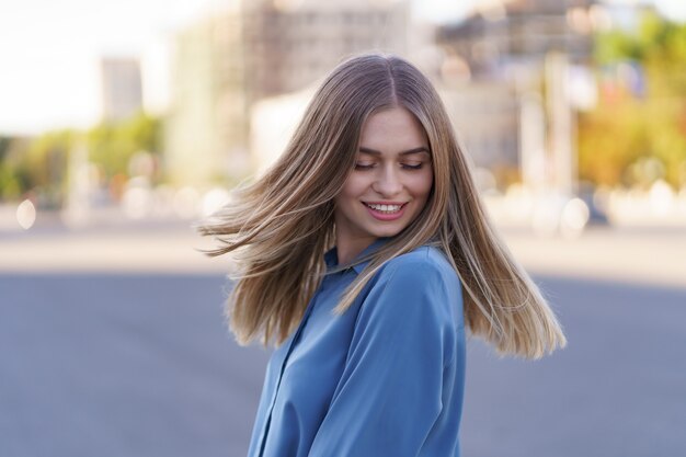 Jolie fille blonde joyeuse avec des cheveux longs volants souriant en riant dans la ville