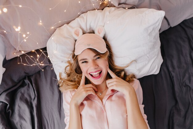 Photo gratuite jolie fille blonde en masque mignon s'amusant tôt le matin. superbe modèle féminin bouclé couché dans son lit et souriant.