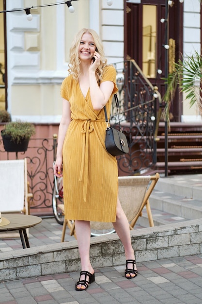 Jolie fille blonde souriante en robe parlant joyeusement sur téléphone portable dans la rue de la ville