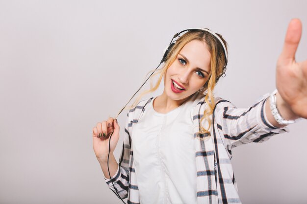 Jolie fille blonde en vêtements blancs s'amusant et dansant drôle. Superbe jeune femme aux cheveux bouclés écoutant la musique préférée dans les écouteurs.