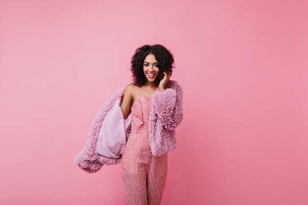 Jolie fille bouclée à la peau foncée pose joyeusement pour sa séance photo de printemps. Portrait de modèle souriant en tenue délicate de couleur poudrée.