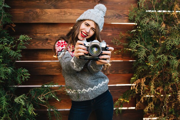 Jolie fille brune aux cheveux longs en vêtements d'hiver s'amuser avec la caméra sur les branches vertes entourent en bois.