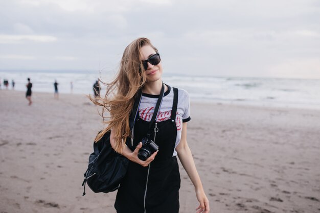 Jolie fille brune avec caméra posant sur la nature en journée nuageuse. Plan extérieur d'une belle dame avec un sac à dos noir se promenant sur la plage.