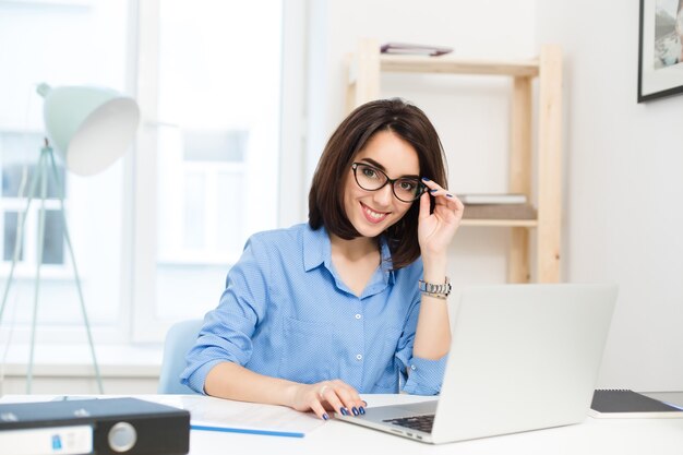 Une jolie fille brune en chemise bleue est assise à la table au bureau. Elle travaille avec un ordinateur portable. Elle sourit à la caméra.