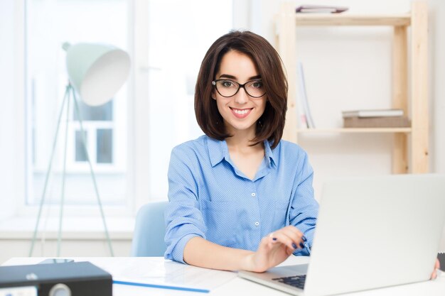 Une jolie fille brune en chemise bleue est assise à la table au bureau. Elle travaille avec un ordinateur portable et sourit à la caméra.