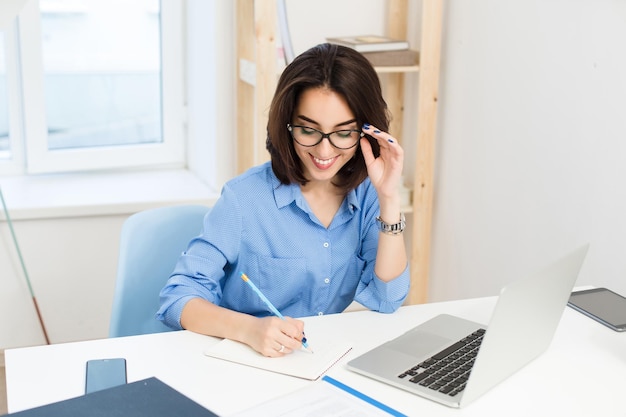 Une jolie fille brune en chemise bleue travaille à table au bureau. Elle écrit dans un cahier avec un sourire.